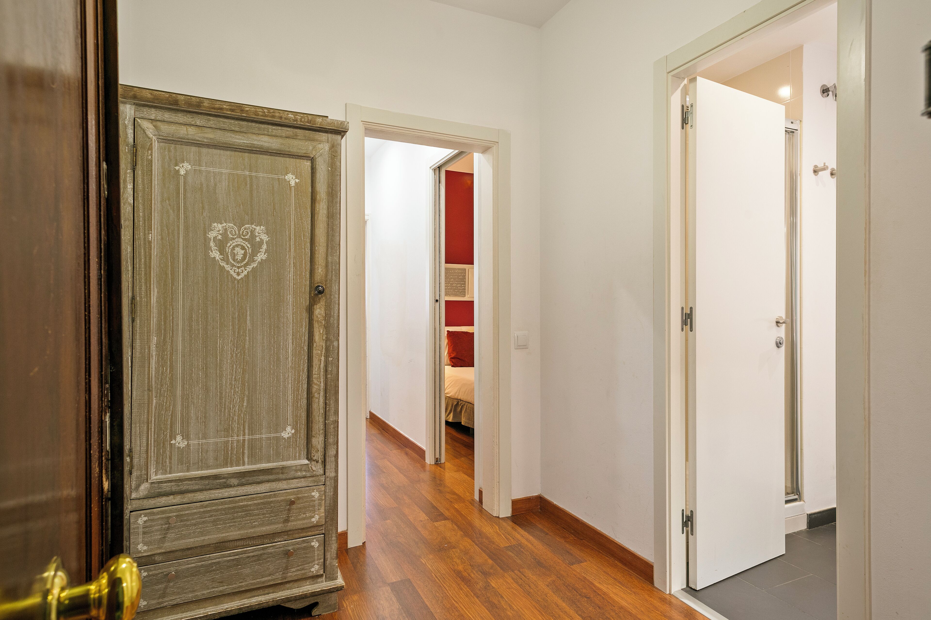 A hallway with wooden flooring, a vintage wardrobe, and a door leading to a room with a bed and red walls.