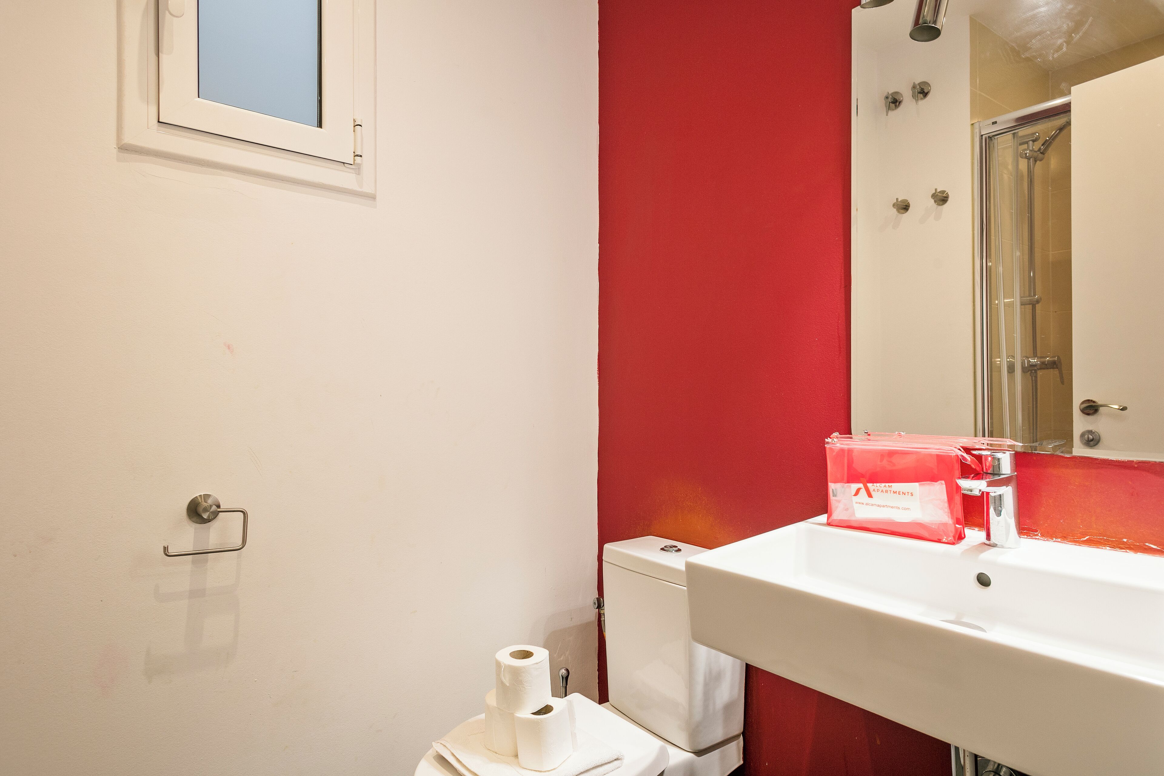 Un baño con lavamanos blanco, pared de acento rojo, inodoro con papel higiénico y zona de ducha.