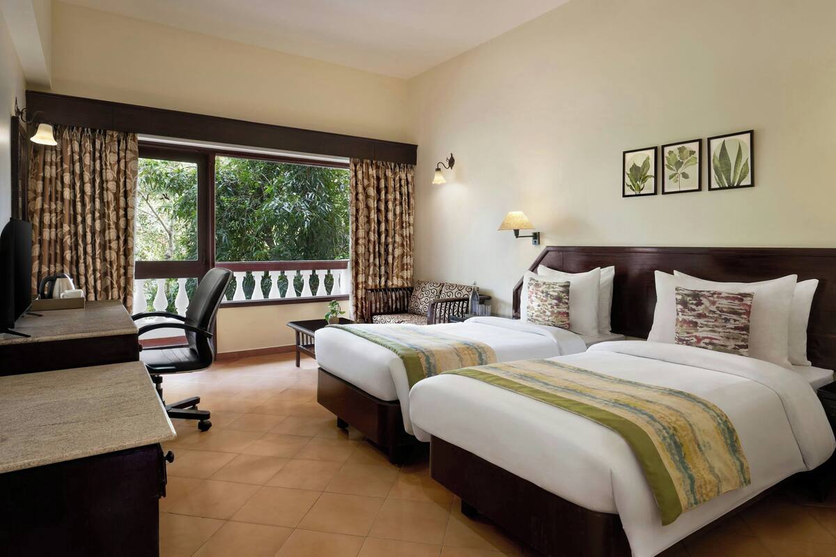 A hotel room with two beds, a desk, a chair, and a balcony with curtains.