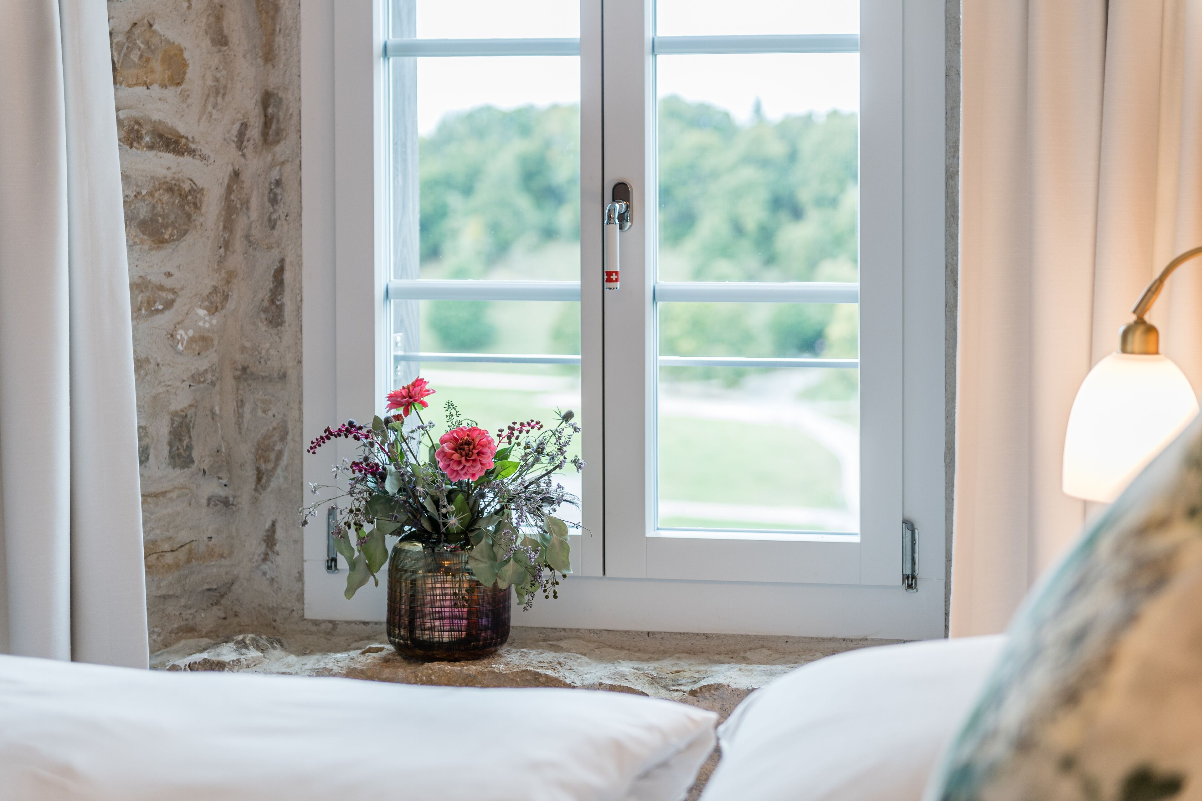 Ein Zimmer mit einem Bett, einem Fenster mit Blick ins Grüne und einer Vase mit Blumen auf der Fensterbank.