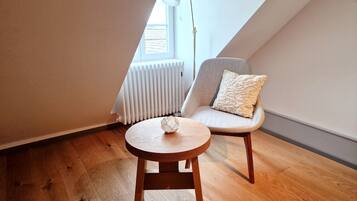 A cozy room with a wooden floor, a round wooden table, a grey armchair with a decorative pillow, and a white radiator.