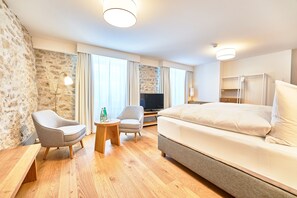 A hotel room with a large bed, two armchairs, a small table, a TV, and a minibar. - Hotel Weissenstein (Oberdorf)