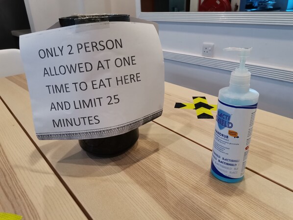 Una mesa con un cartel que indica 'SOLO DOS PERSONAS PUEDEN COMER AQUÍ A LA VEZ Y TIEMPO LÍMITE DE 25 MINUTOS' y una botella de Bactishield desinfectante para manos.