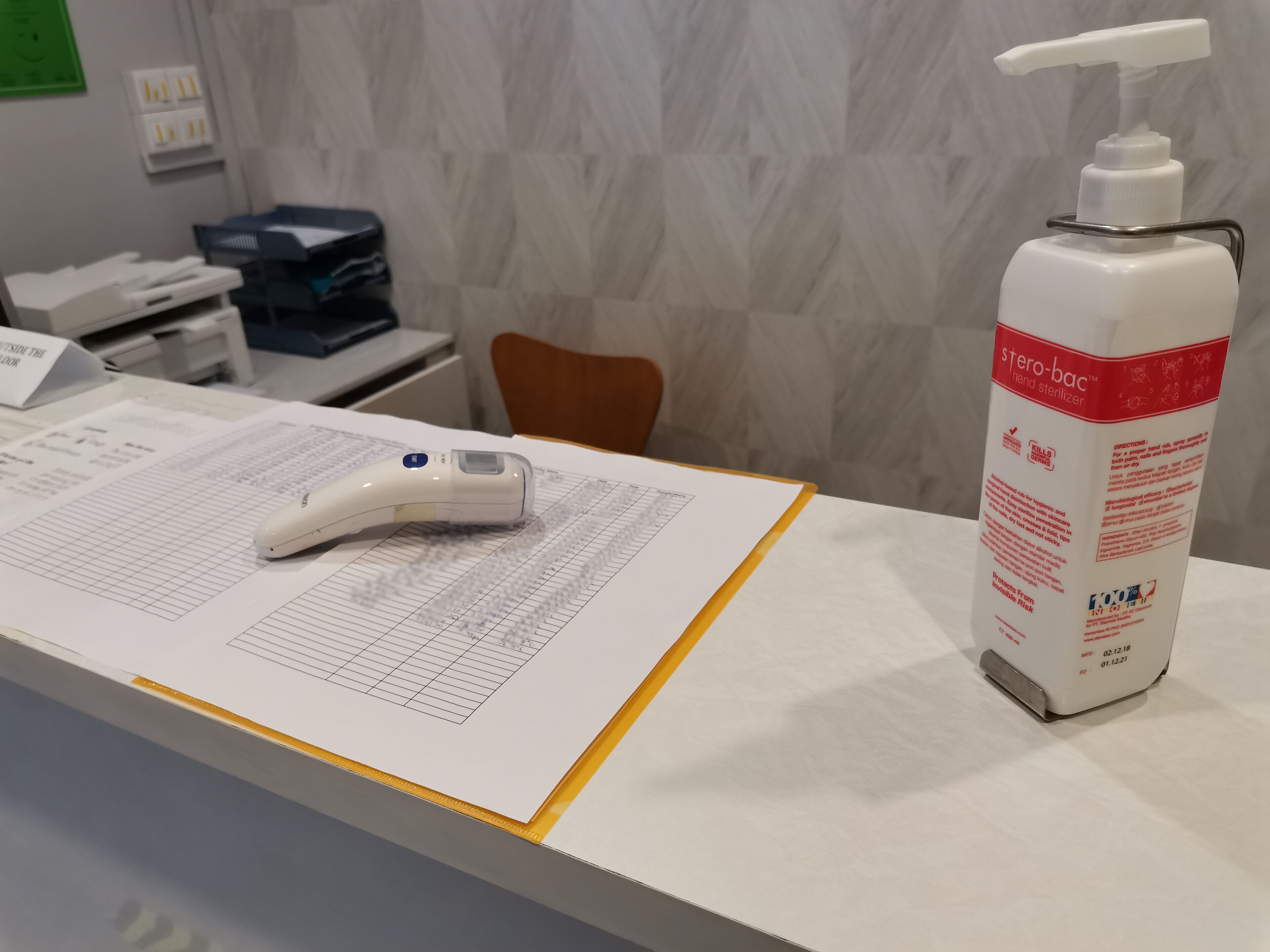 En receptionsdisk med en handspritdispenser, en digital termometer och en skrivtavla med dokument.