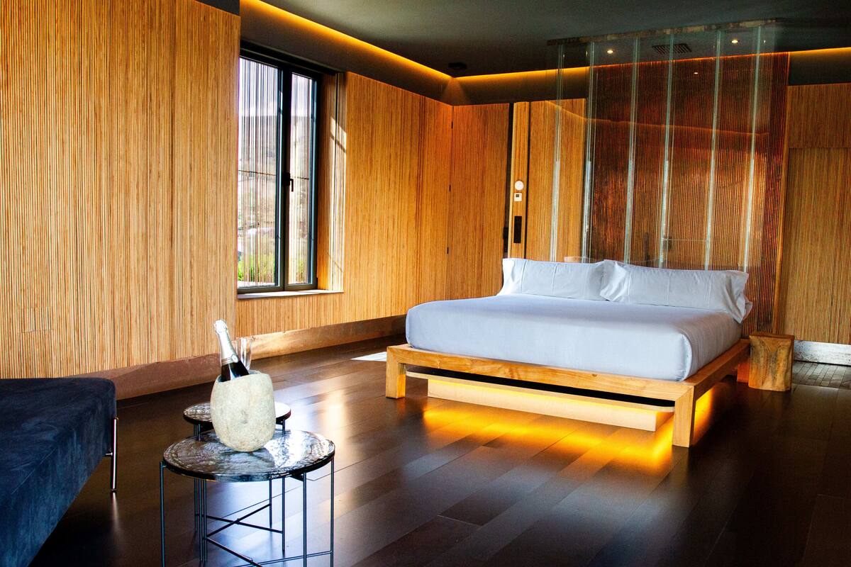 A modern bedroom with a large bed, wooden walls, a small round table, and a window.