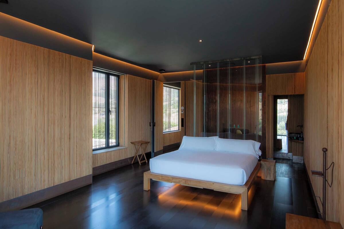 A modern bedroom with a large bed, wooden panel walls, and large windows.