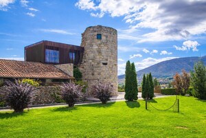 A stone tower adjacent to a modern building with a tiled roof, surrounded by a well-maintained lawn and cypress trees.