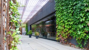 A covered outdoor corridor with potted plants, a glass wall, and a metal fence.