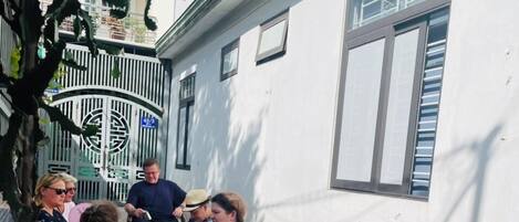 Eine Gruppe von Personen sitzt auf Außenbänken in einem Innenhof mit Fliesenböden. Im Hintergrund ist ein weißes Gebäude mit Fenstern zu sehen.