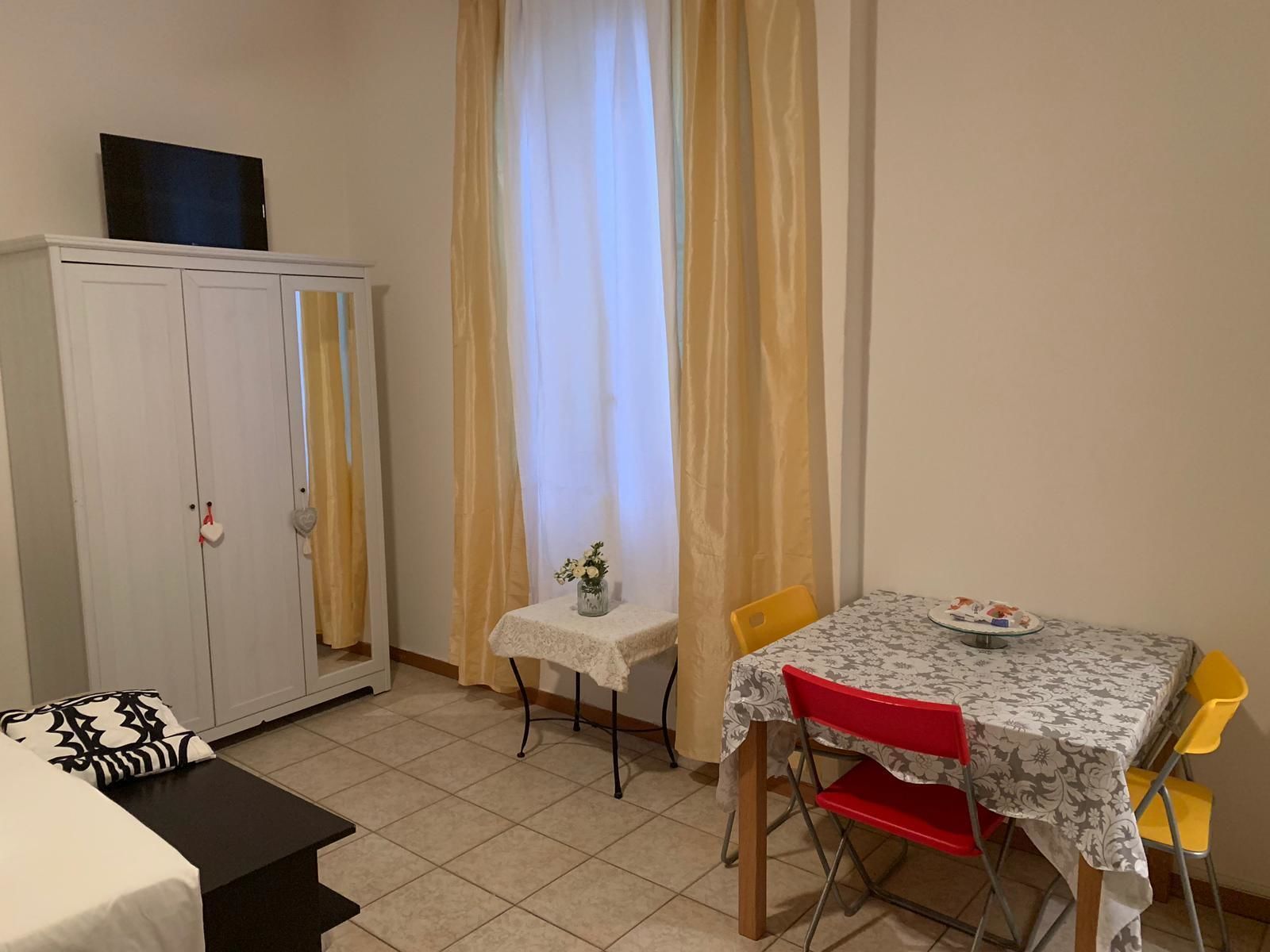 Una habitación pequeña y ordenada con cama, armario, mesa con sillas y ventana con cortinas.