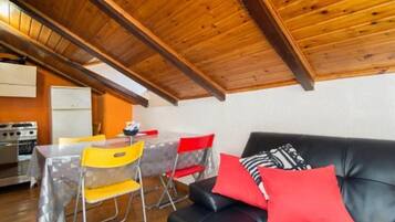 Una habitación acogedora con techo de madera, un sofá de cuero negro con cojines rojos, una mesa de comedor con sillas y una zona de cocina con estufa.