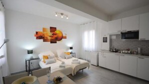 Een moderne, goed verlichte woonkamer met een bank en een eettafel, een keuken met witte kastjes, en een muur met een kleurrijk abstract schilderij.
