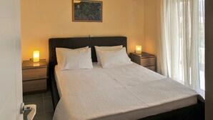 Ein ordentlich bezogenes Bett mit weißen Leinen, zwei Nachttische mit Lampen, ein Bild an der Wand und ein Fenster mit Vorhängen.