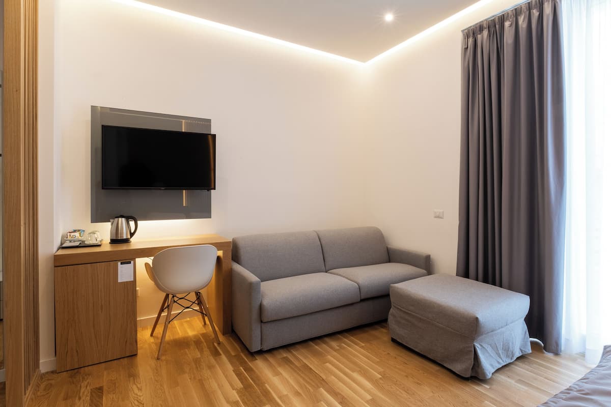 A modern, well-lit room with a flat-screen TV mounted on the wall, a grey sofa, a small ottoman, a wooden desk with a kettle, and a chair.