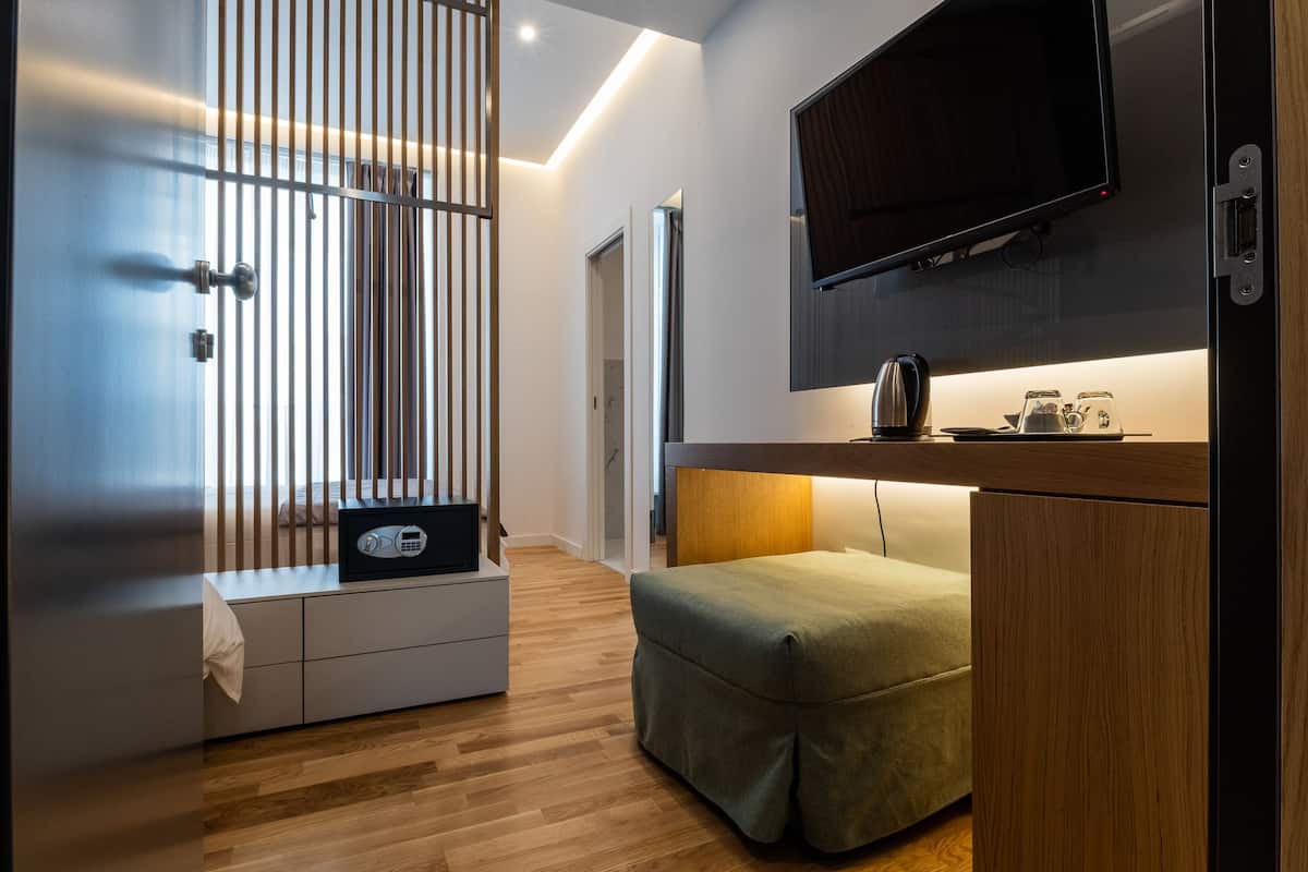 A modern hotel room with a wooden floor, a green upholstered bench, a flat-screen TV mounted on the wall, a small table with a kettle, and a sliding door with vertical blinds.