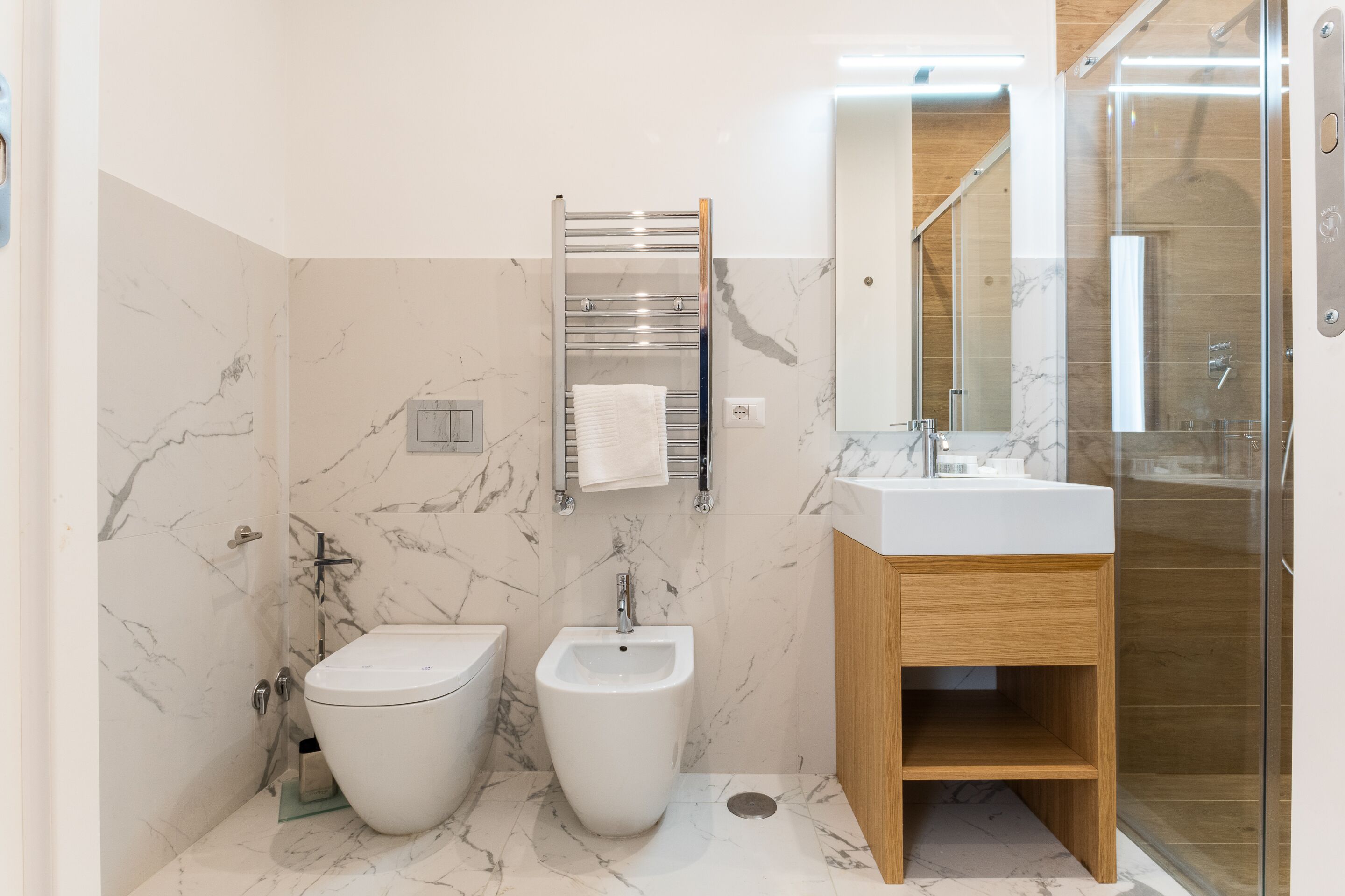 A modern bathroom with a bidet, toilet, and sink, marble walls, and a glass shower enclosure.