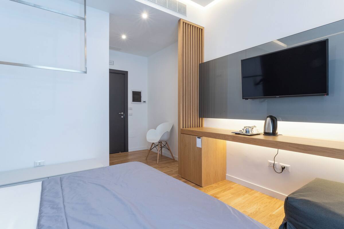 A modern hotel room with a bed, a TV mounted on the wall, a wooden desk, and a chair.
