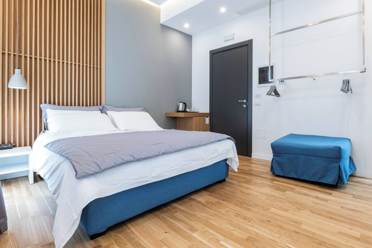 A modern bedroom with a large bed, a wooden slat wall, a bedside table with a lamp, a bench, and a wardrobe with a mirror.