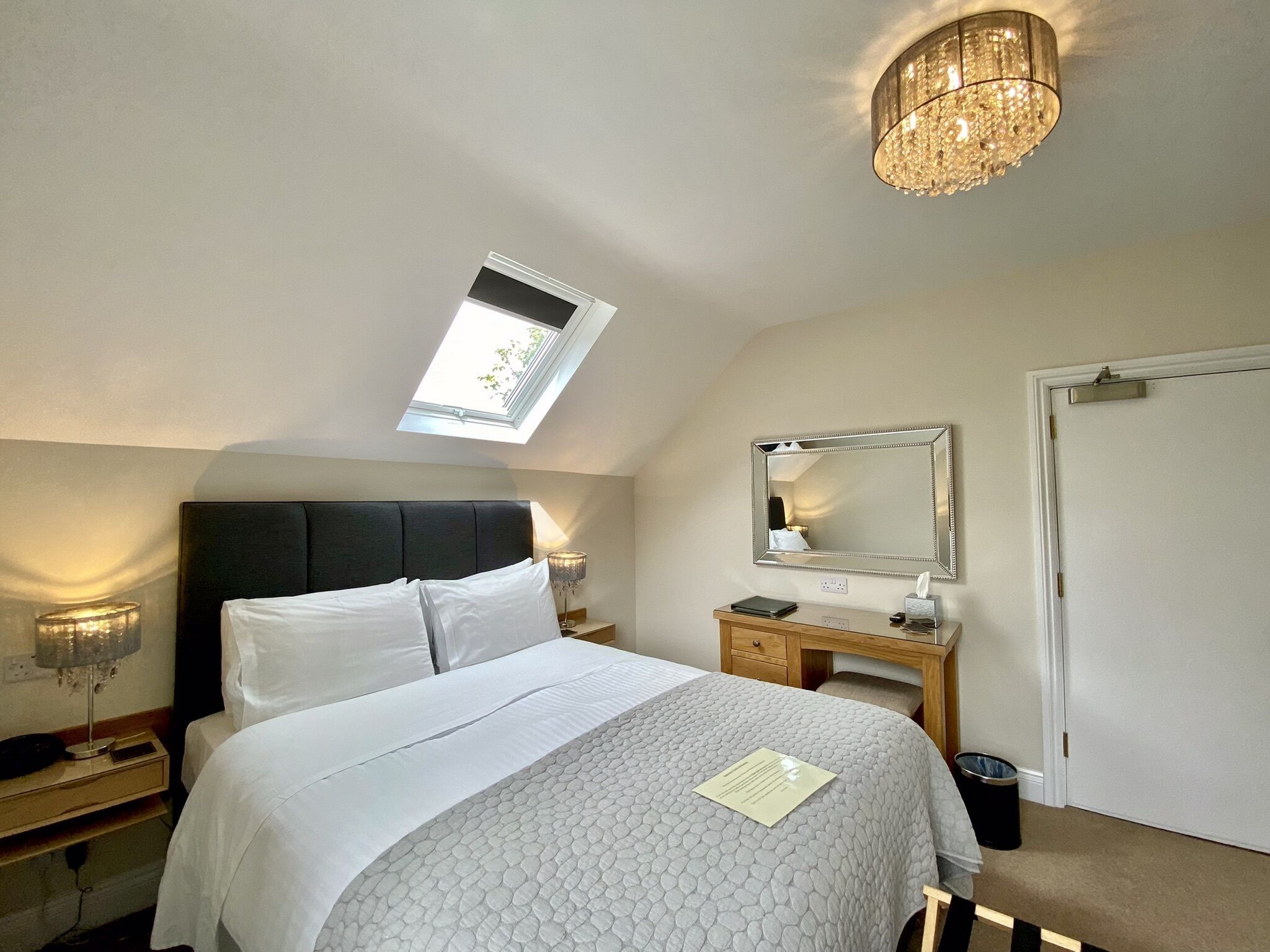 A bedroom with a bed, a bedside table, a nightstand, a mirror, a chandelier, a skylight, and a door.