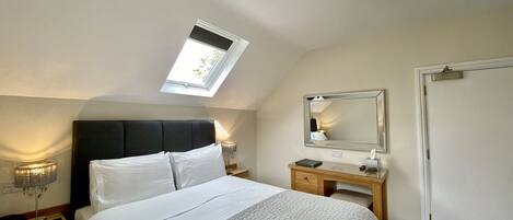 A bedroom with a bed, a bedside table, a nightstand, a mirror, a chandelier, a skylight, and a door.
