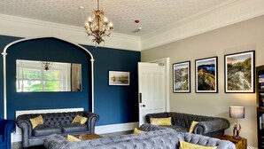A room with a large grey sectional sofa, a smaller sofa, and a blue armchair. There is a chandelier, a fireplace, and framed pictures on the wall. - Crow How Country Guest House (Ambleside)