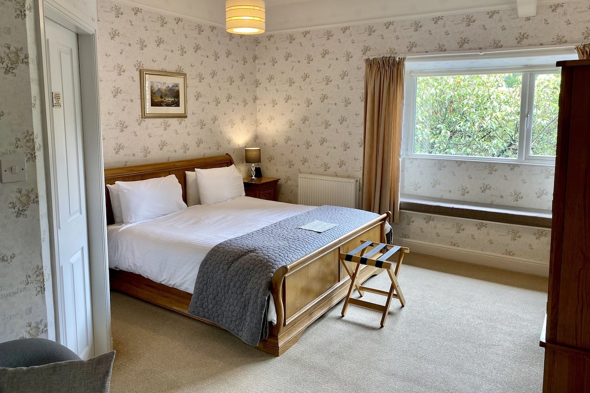 Un lit bien fait, avec du linge de lit blanc et une couverture grise, une table de chevet avec une lampe, une fenêtre avec des rideaux et un banc en bois.