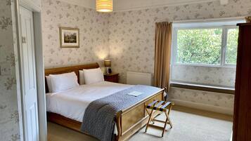 A neatly made bed with white linens and a gray blanket, a bedside table with a lamp, a window with curtains, and a wooden bench.