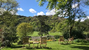 A grassy outdoor seating area with wooden chairs and tables, surrounded by trees and a view of a green valley with hills in the background. - Crow How Country Guest House (Ambleside)