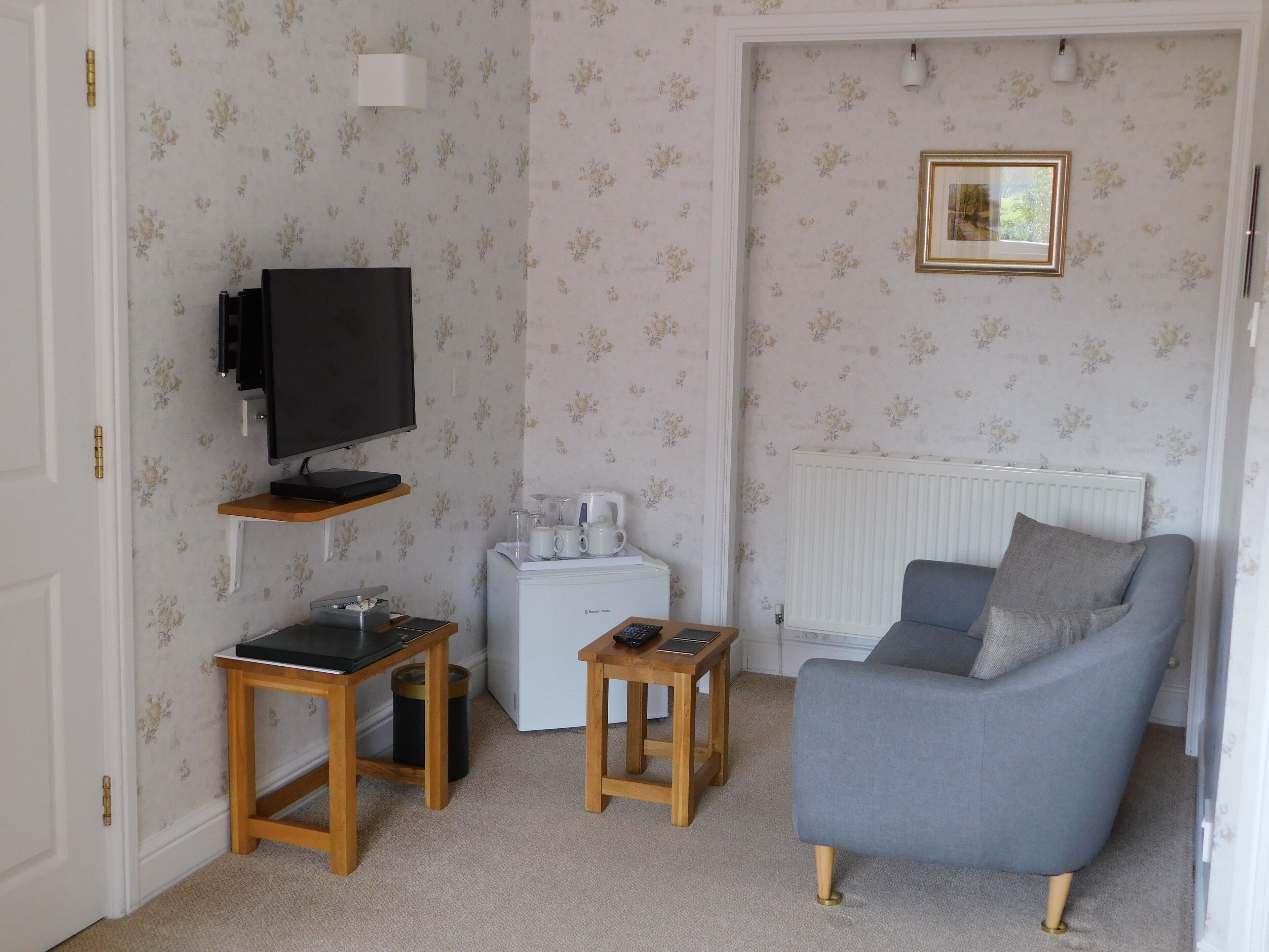 A living room with a TV, two wooden tables, a sofa, a radiator, and a small coffee station.