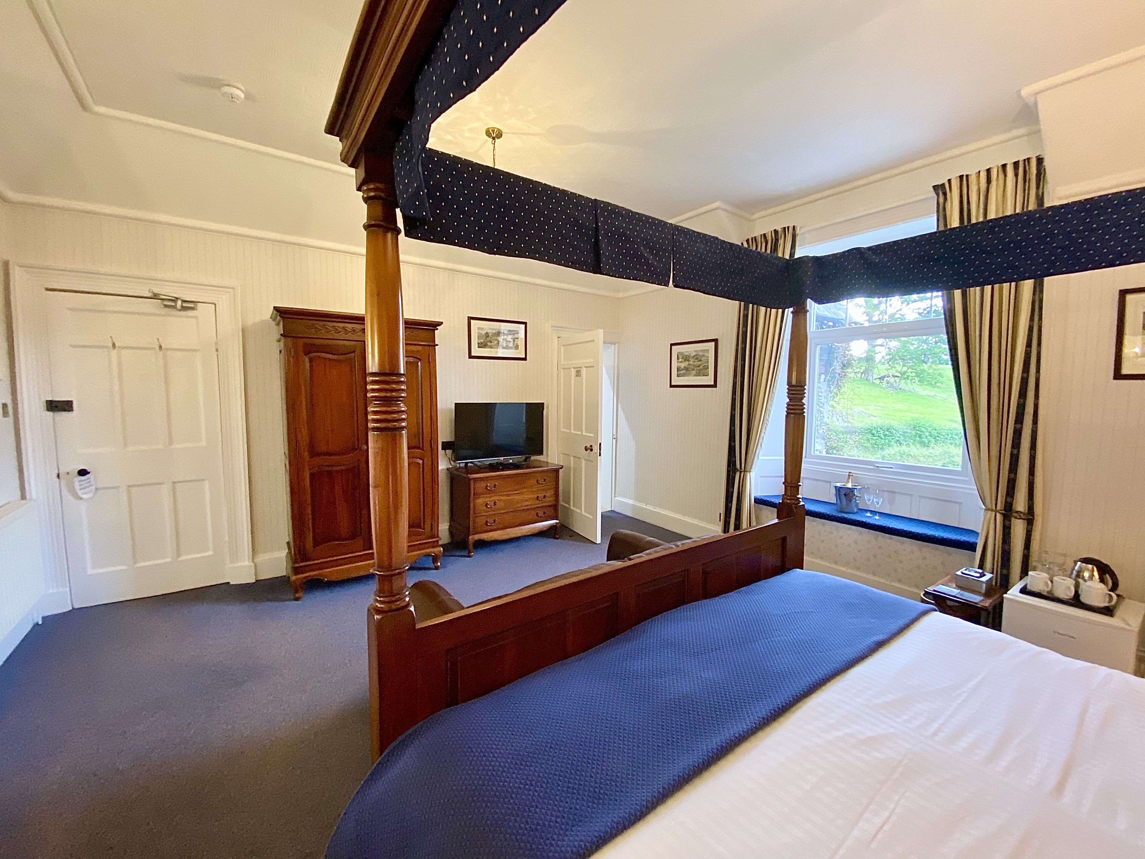 A four-poster bed with a blue bedspread, a wooden dresser, a television on a stand, and a window with curtains.