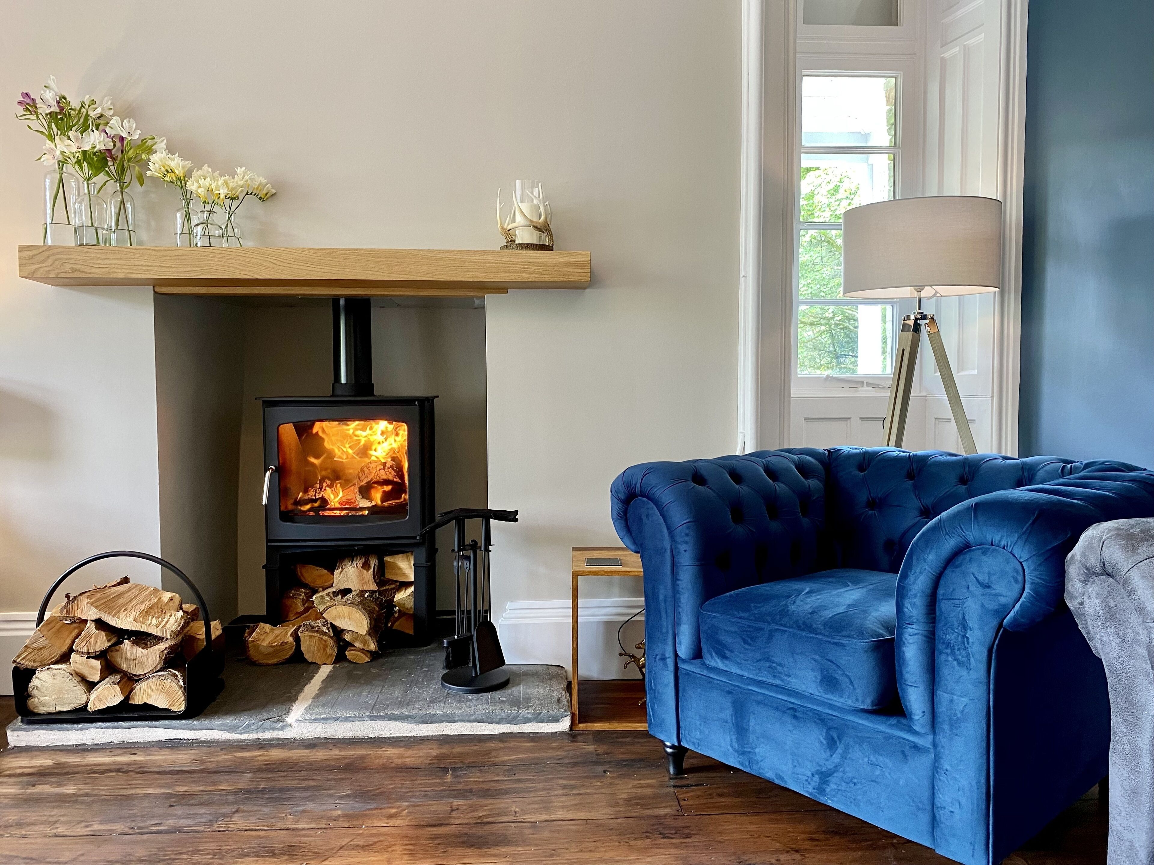 Un salon chaleureux avec un poêle à bois, un canapé en velours bleu et une étagère en bois ornée de fleurs.