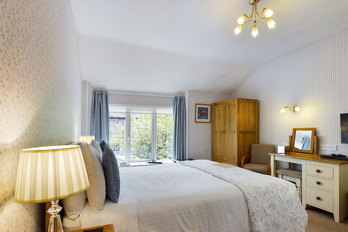 Une chambre à coucher comprenant un lit, une table de chevet, une lampe, une armoire, un bureau, une chaise et une fenêtre avec des rideaux.