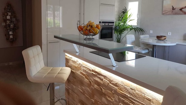 A modern kitchen with white cabinets, a central island with a stone accent, and a dining area with a white chair.