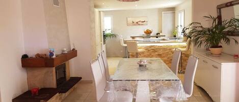 A dining area with a glass table, white chairs, a fireplace, and a kitchen in the background.