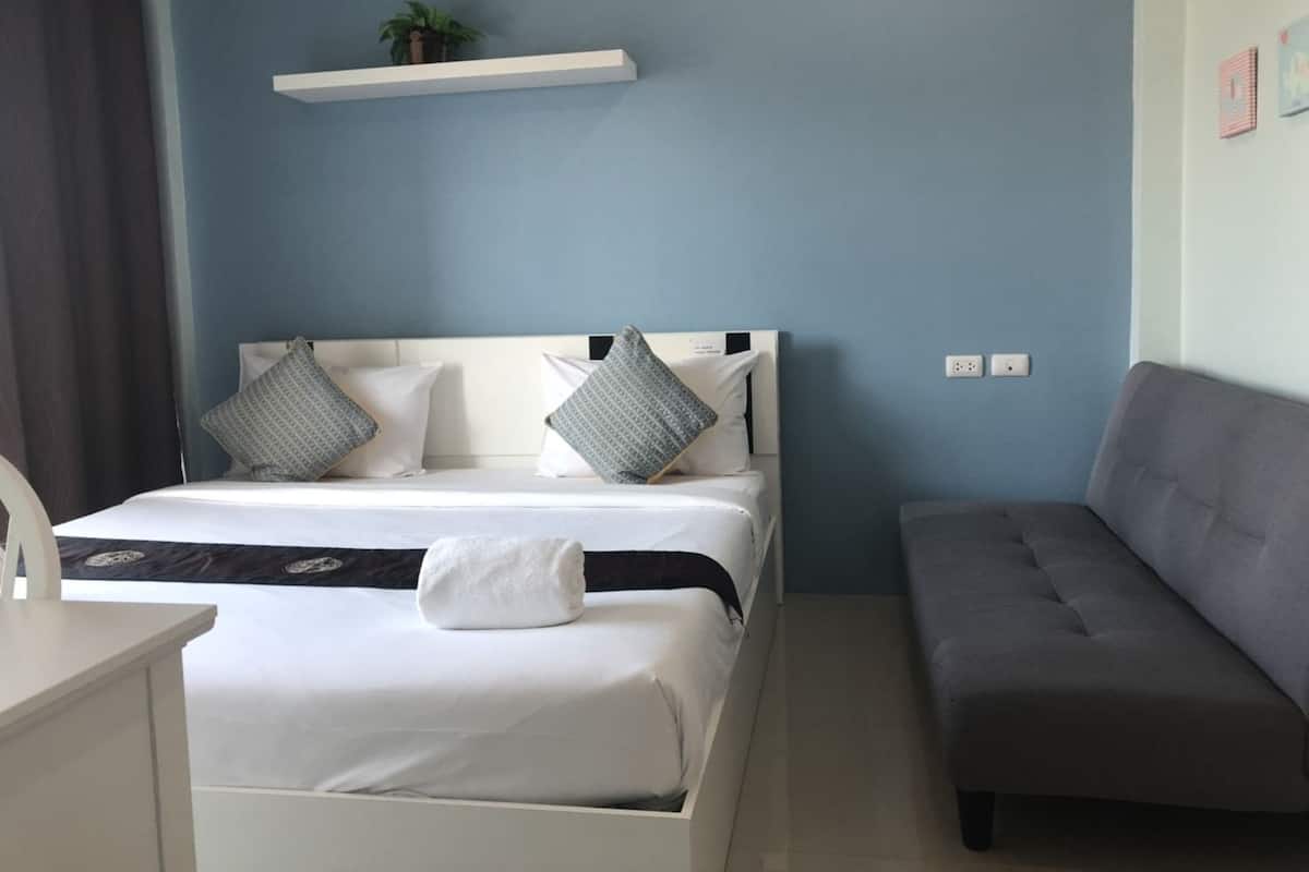 Een slaapkamer met een bed, een nachtkastje, een grijze bank, een wandplank met een plant en een stopcontact.