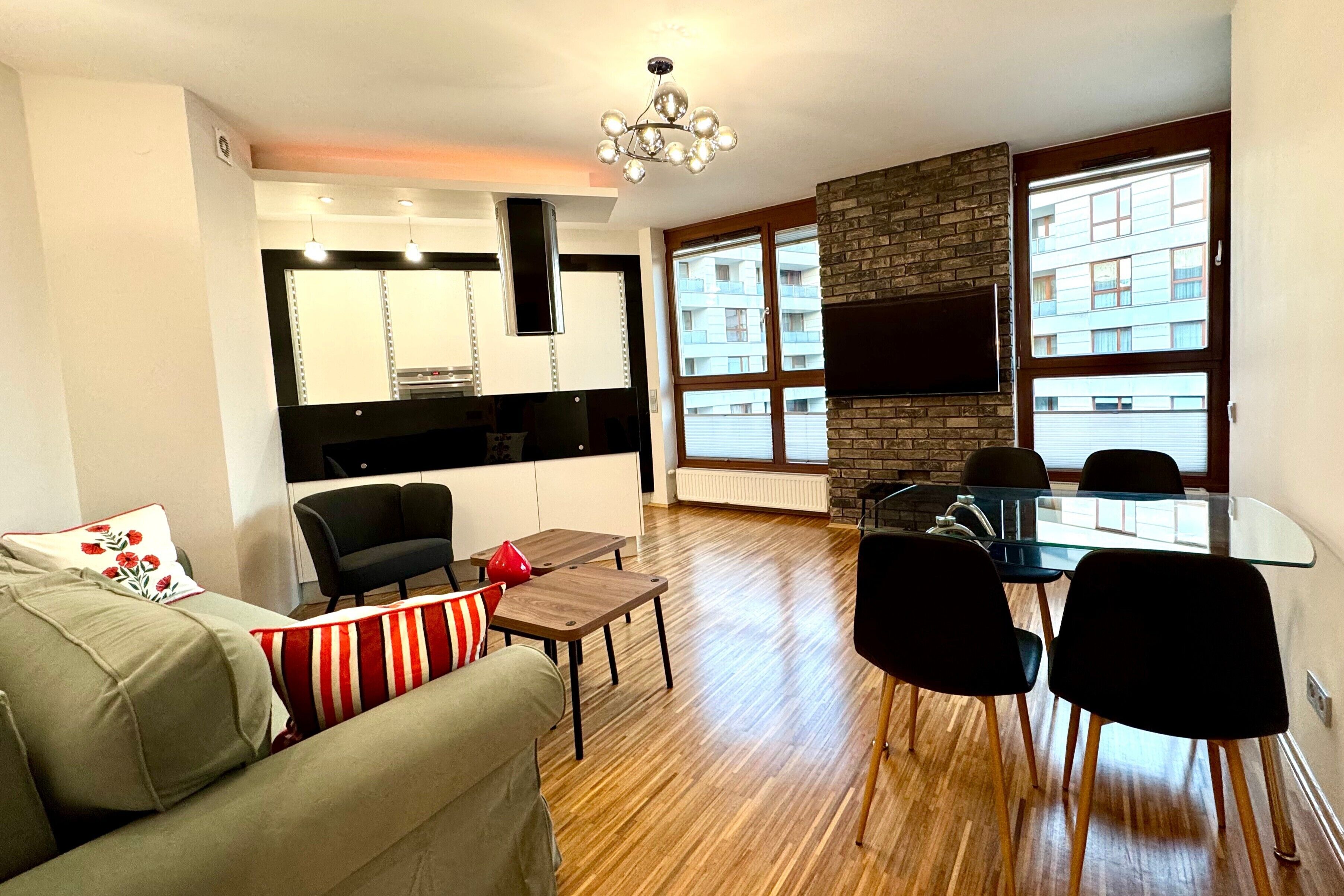 A modern living room with a sofa, a glass dining table, and black chairs. There is a kitchen area with a black countertop and a built-in oven. A large window provides a view of a building outside.