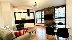 Un salon moderne avec un canapé, une table à manger en verre et des chaises noires. On y trouve également un coin cuisine avec un plan de travail noir et un four encastré. Une grande fenêtre offre une vue sur un bâtiment situé à l’extérieur.