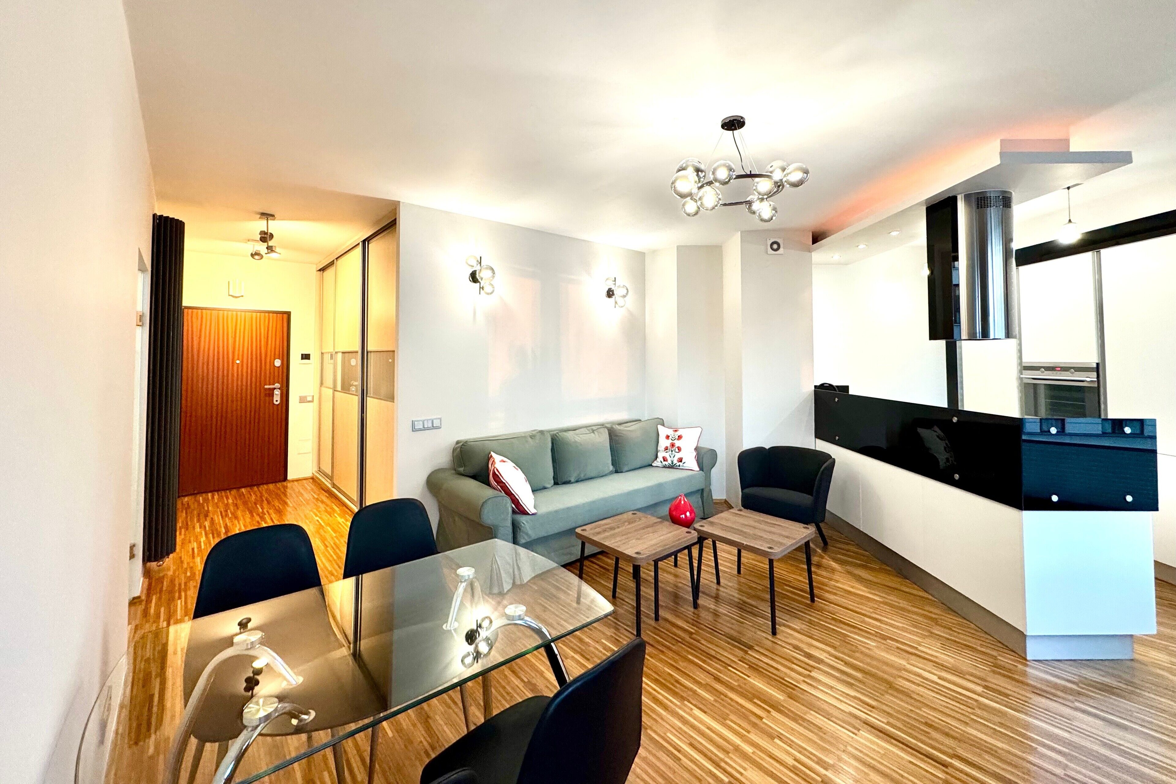 A modern living room with a glass dining table, a sofa, and a kitchen area with a stove.