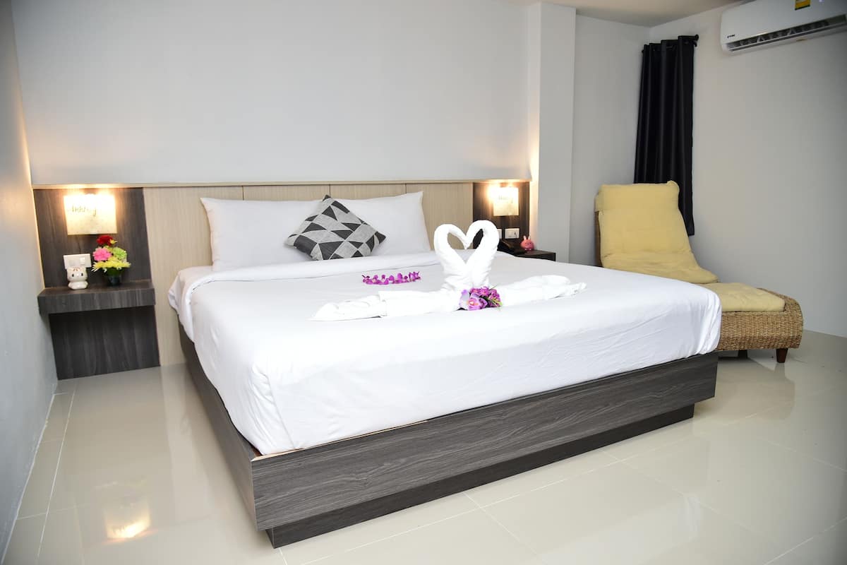 Cama bem arrumada com roupa de cama branca, arranjo de toalhas em formato de coração, travesseiro geométrico e uma cadeira amarela ao lado.