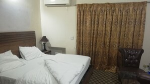 A hotel room with a bed, a chair, an air conditioning unit, a lamp, and a patterned curtain. - Greenland Hotel (Lahore)