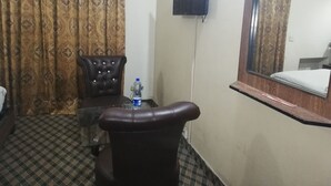 A hotel room with a checkered floor, a brown leather chair, a small table with a water bottle, a flat-screen TV mounted on the wall, and a large mirror above a bed. - Greenland Hotel (Lahore)