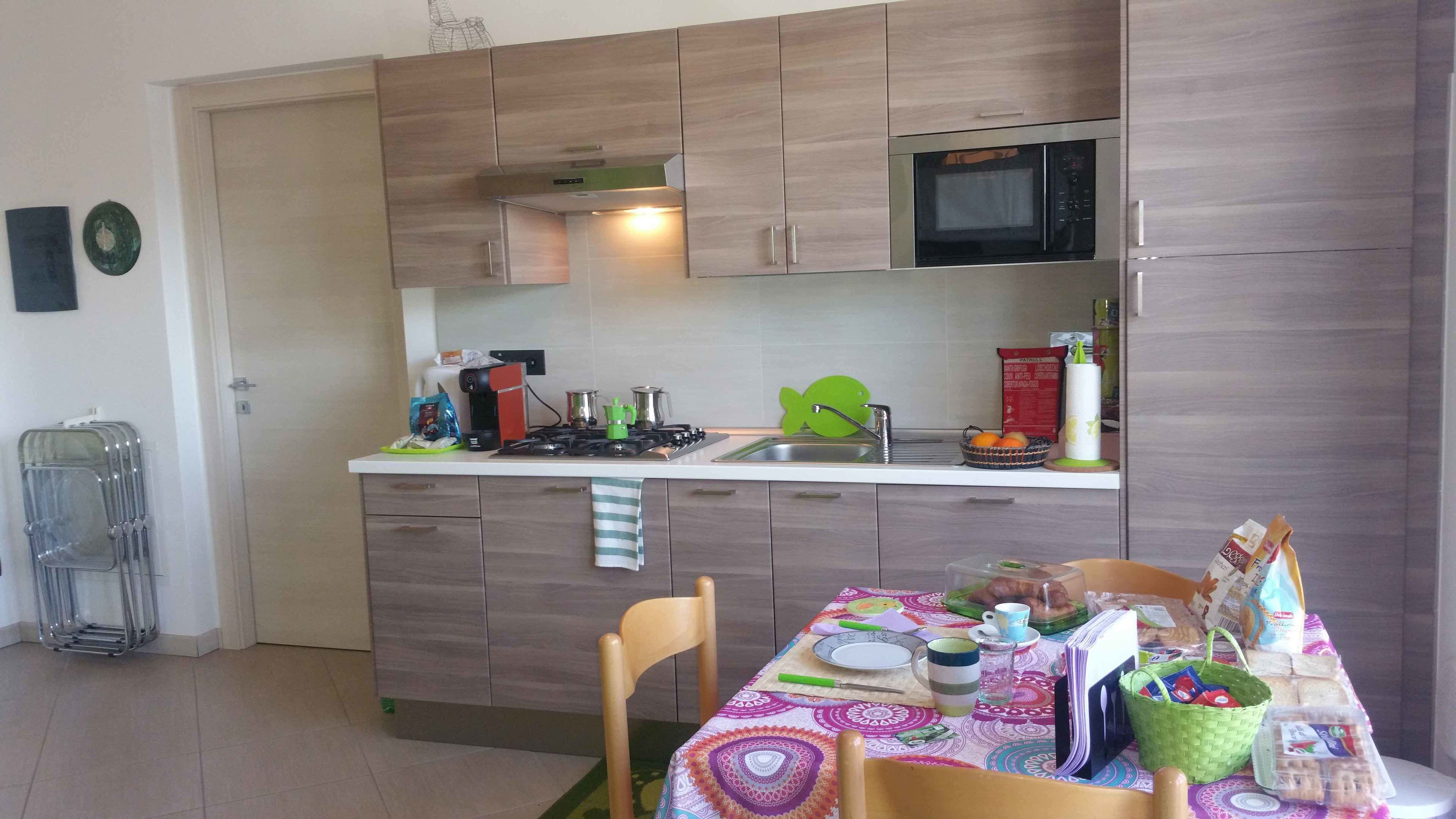 A compact kitchen with wooden cabinets, a microwave, a gas stove, and a sink. There is a dining table with a colorful tablecloth and various food items.