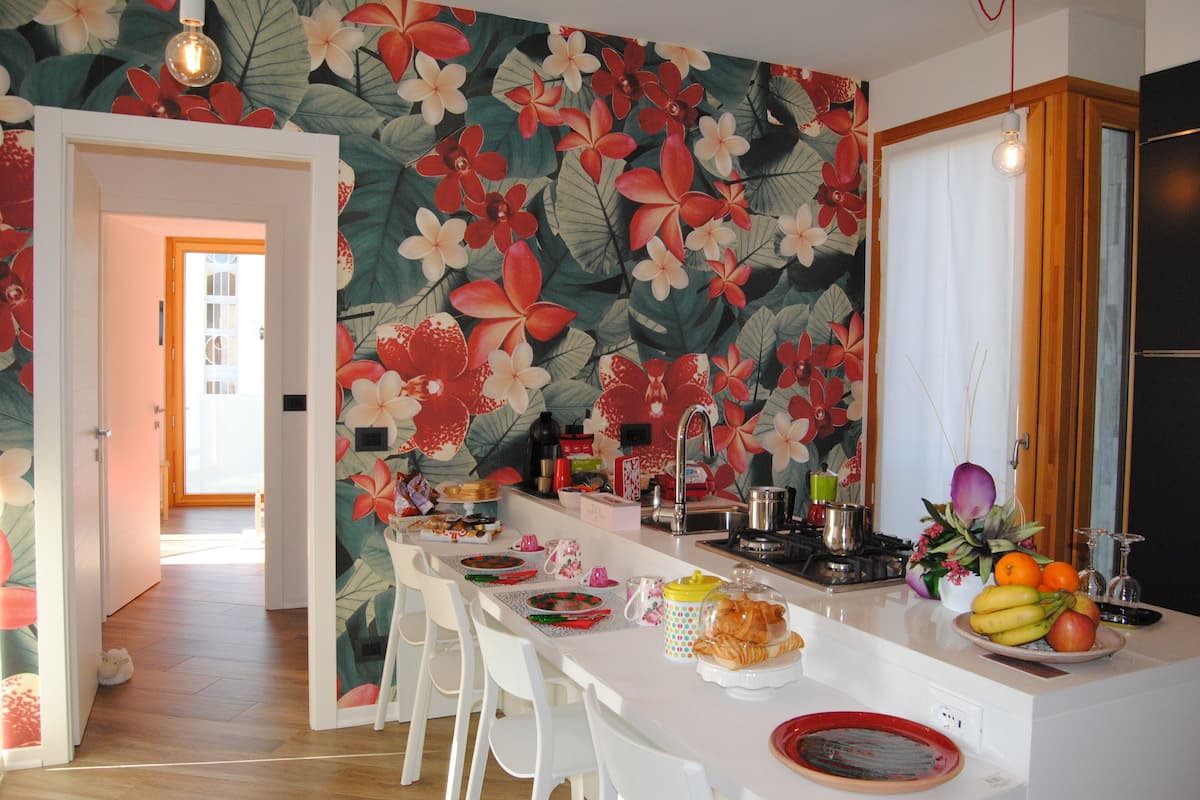 Una cocina con papel tapiz floral, taburetes de bar blancos, un fregadero y un cuenco de frutas.