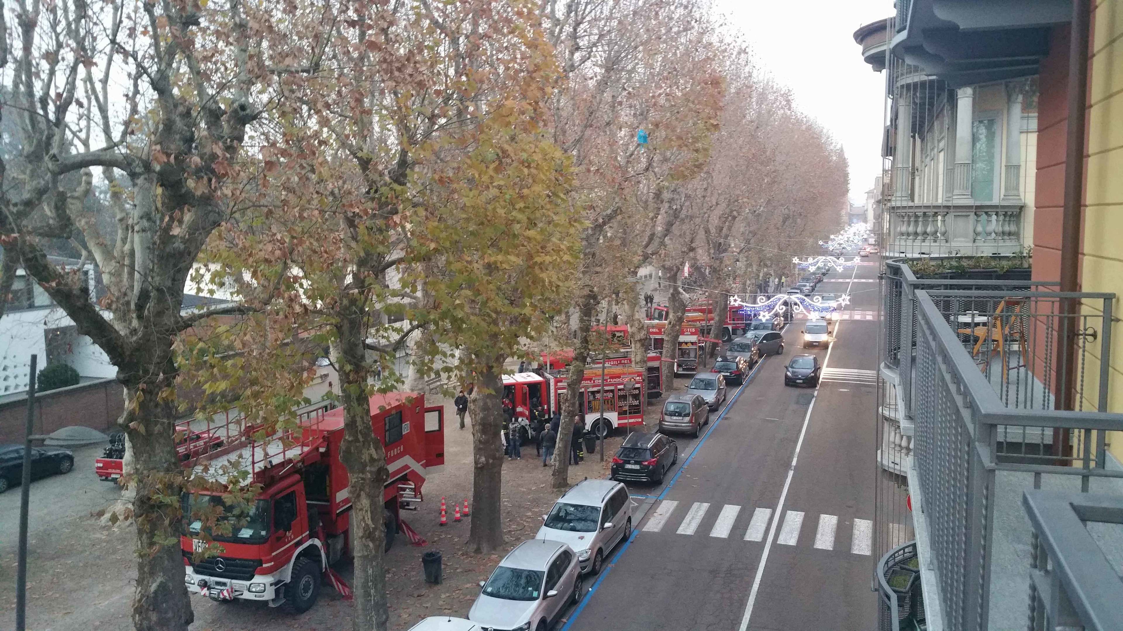 Une rue bordée d’arbres et de voitures garées, où l’on aperçoit un camion de pompiers et du personnel d’intervention d’urgence.