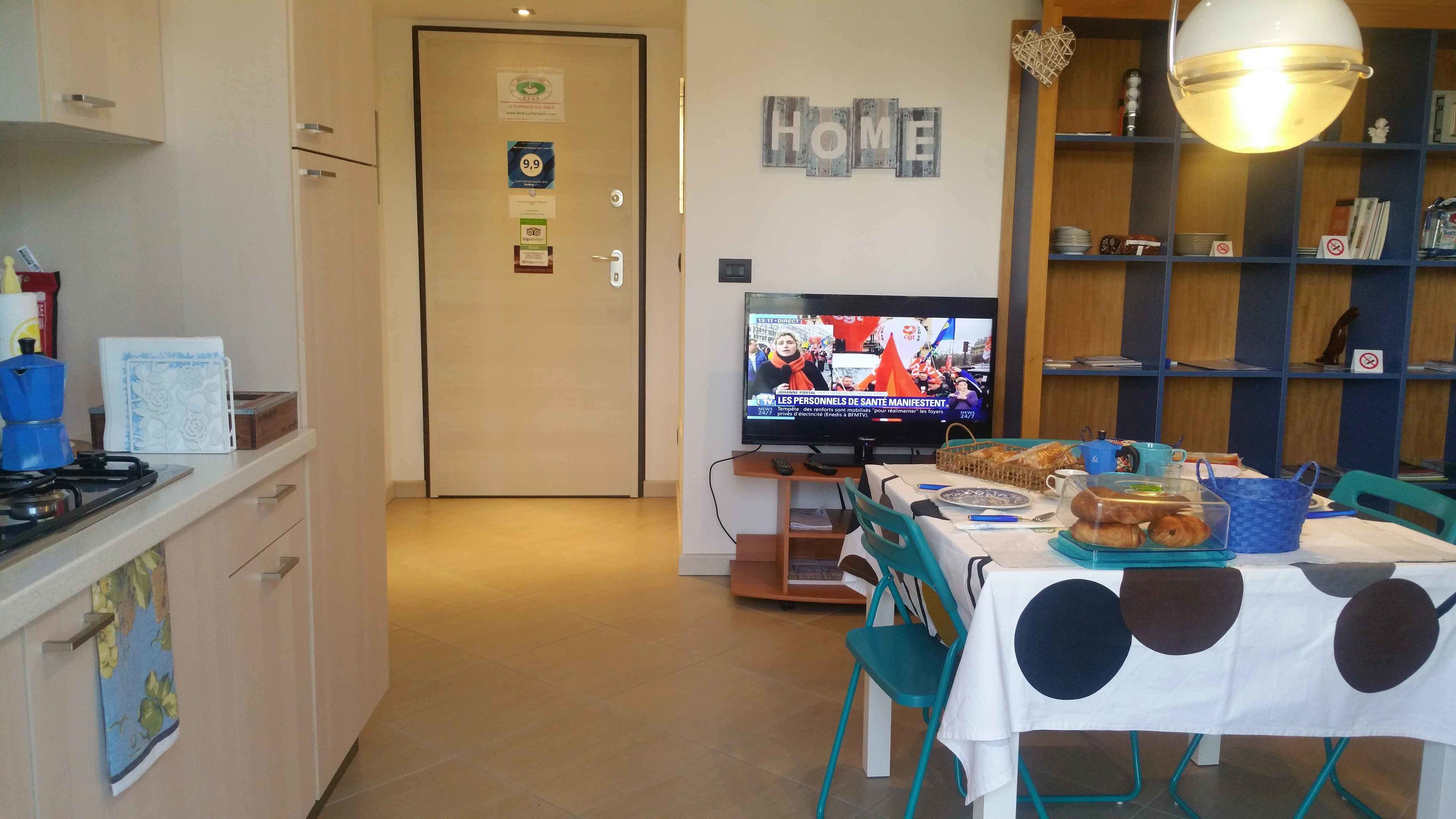A kitchen with a dining area, a television displaying a news broadcast, and a 'HOME' sign on the wall.