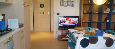A kitchen with a dining area, a television displaying a news broadcast, and a 'HOME' sign on the wall.