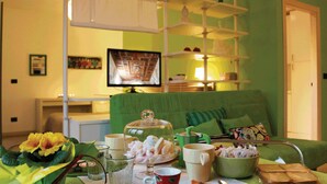 Una habitación con un sofá verde, una mesita con un vaso, un cuenco y una bandeja con galletas. Se observa una estantería con tazas y un televisor al fondo.