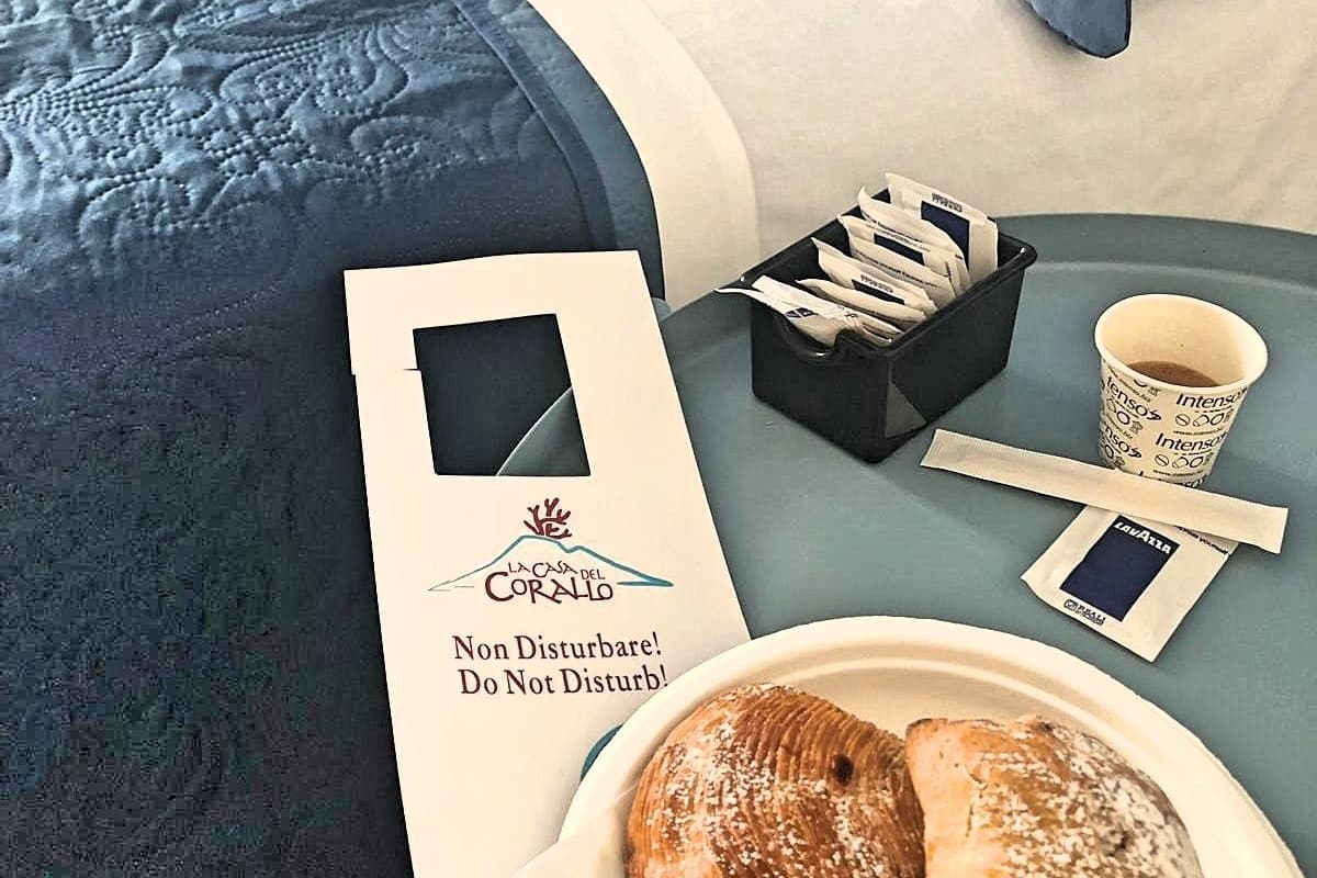 Ein ordentlich bezogenes Bett mit blauer Bettdecke und Kissen, ein Tablett mit Croissants und einer Kaffeetasse, sowie eine Karte mit dem Hotel-Logo.