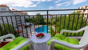A balcony with two white chairs, a table with a plate of strawberries, and a view of a swimming pool and buildings in the background. - Large Studio in Polo Resort (Sunny Beach)
