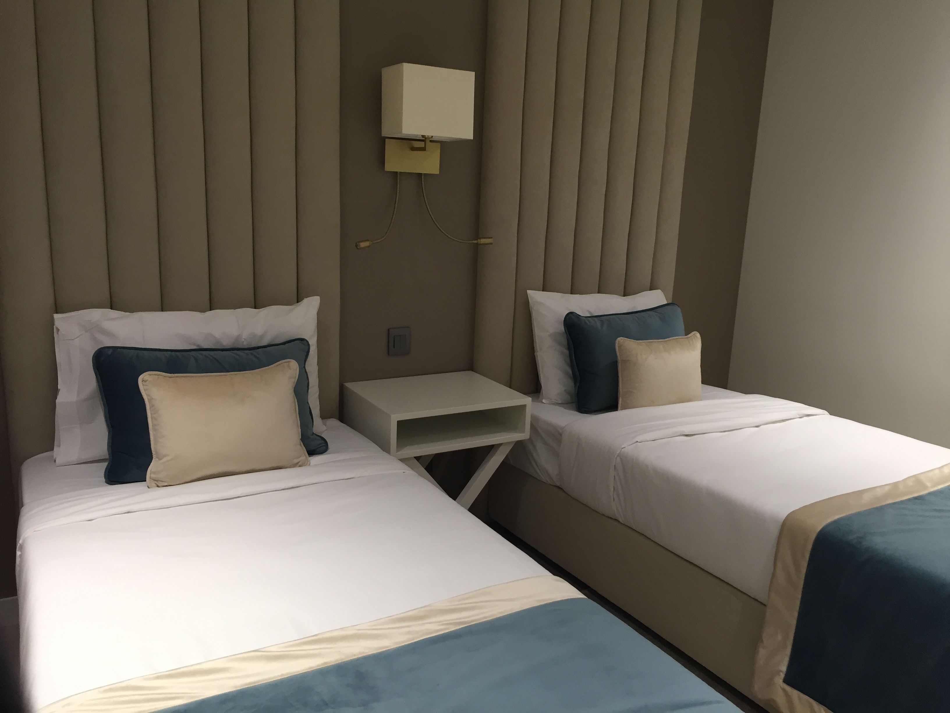 A hotel room with two beds, each with white bedding and blue accents. There is a small bedside table between the beds, and a wall-mounted light fixture above it.